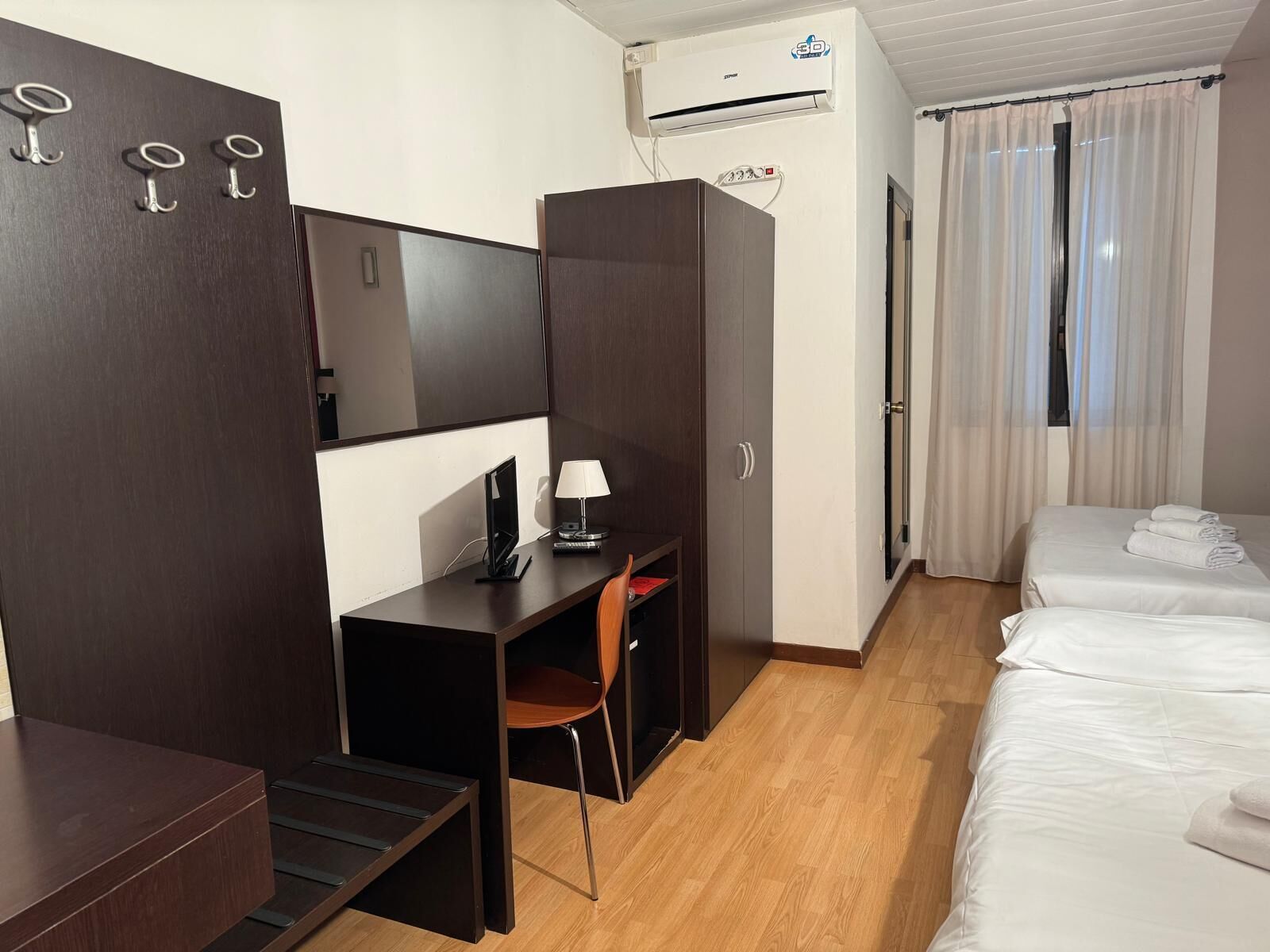 A hotel room with a desk, chair, two beds, and an air conditioning unit.