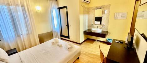 Habitación de hotel con cama, escritorio, silla, baño con lavabo y espejo, y ventana con cortinas.