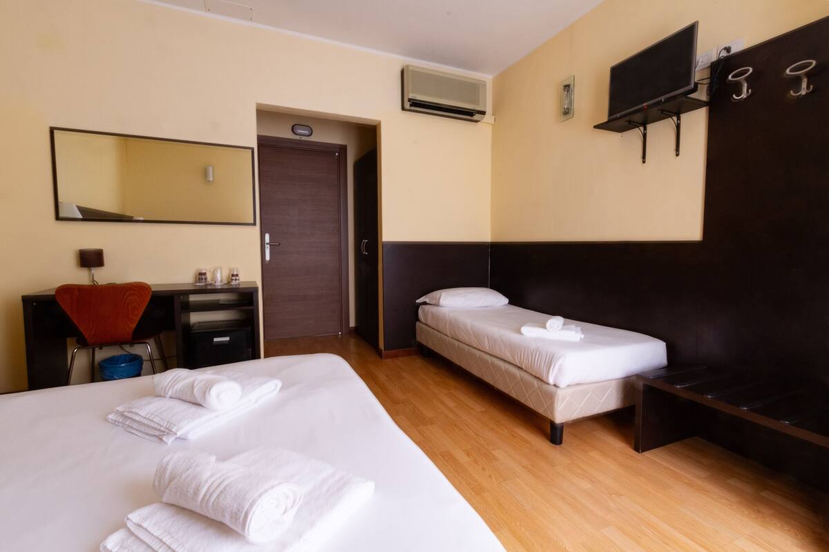 A hotel room with a large bed, a smaller bed, a desk with a chair, a TV mounted on the wall, and an air conditioning unit.