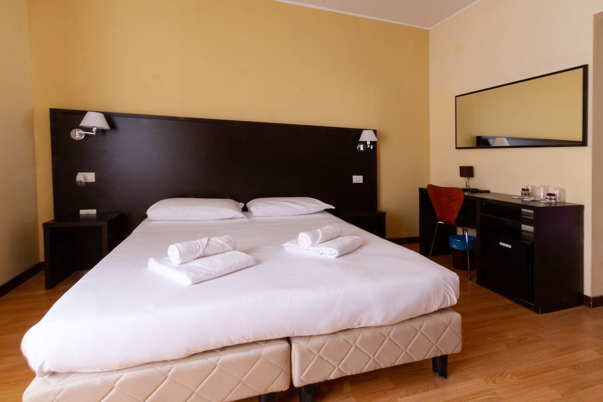 A neatly made bed with white linens and towels, a dark wooden headboard, bedside lamps, a desk with a chair, and a large mirror on the wall.
