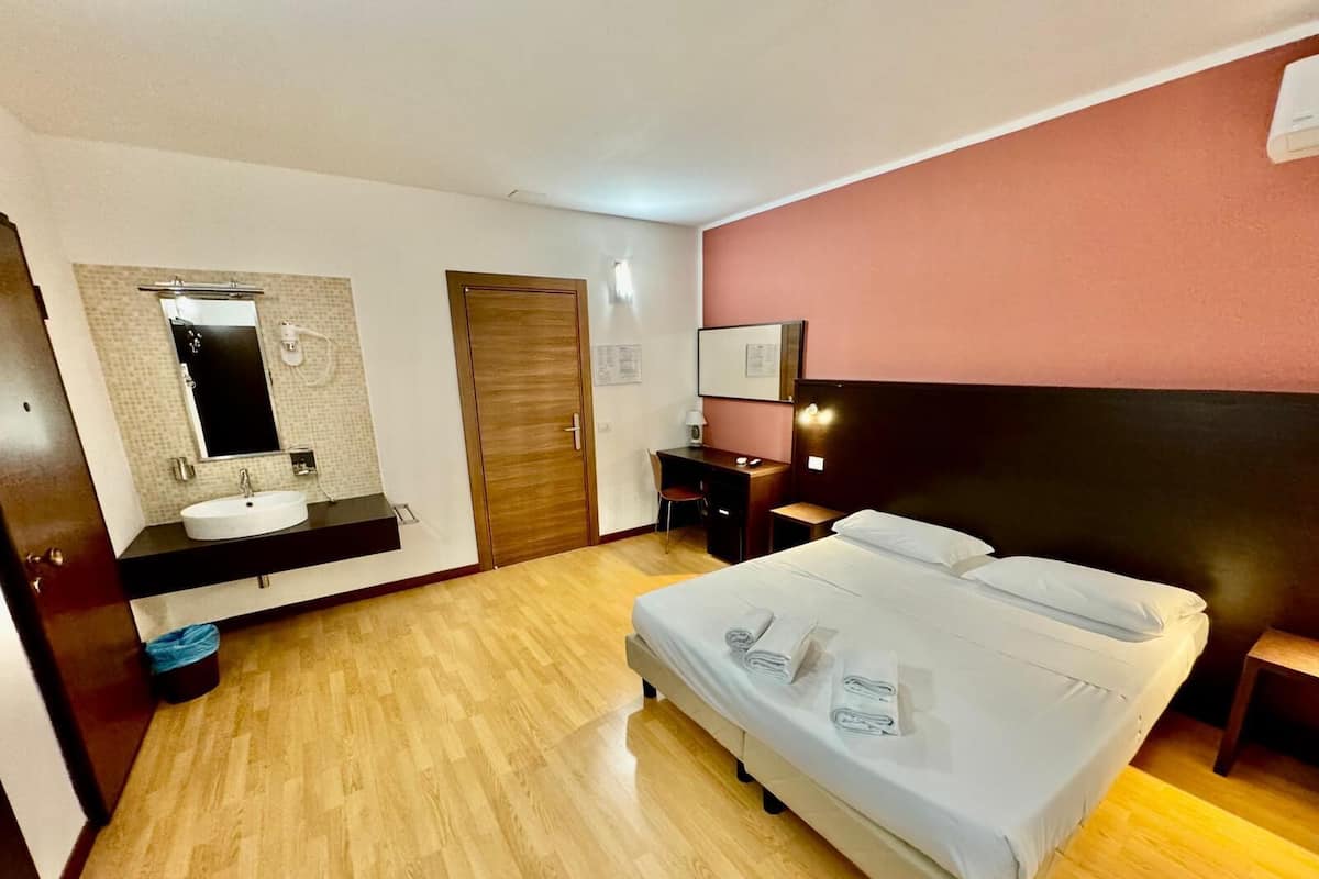 A hotel room with a wooden floor, a bed with white linens, a desk with a chair, a wooden door, and a bathroom with a sink and mirror.