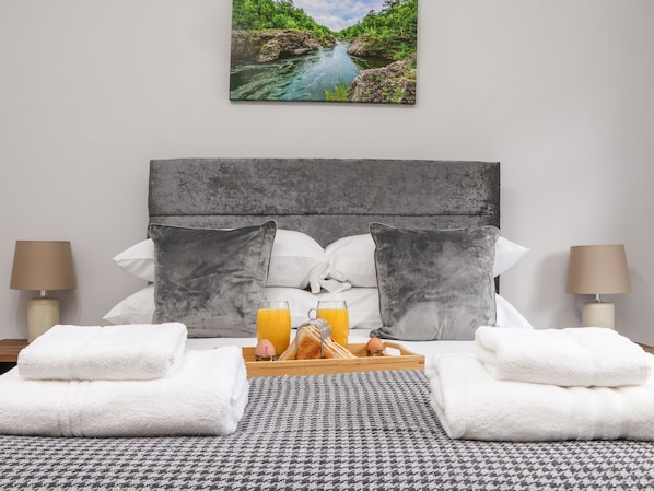 Une chambre d’hôtel avec un lit, deux oreillers, un plateau avec des serviettes et du jus d’orange, et un tableau représentant une rivière accroché au mur.