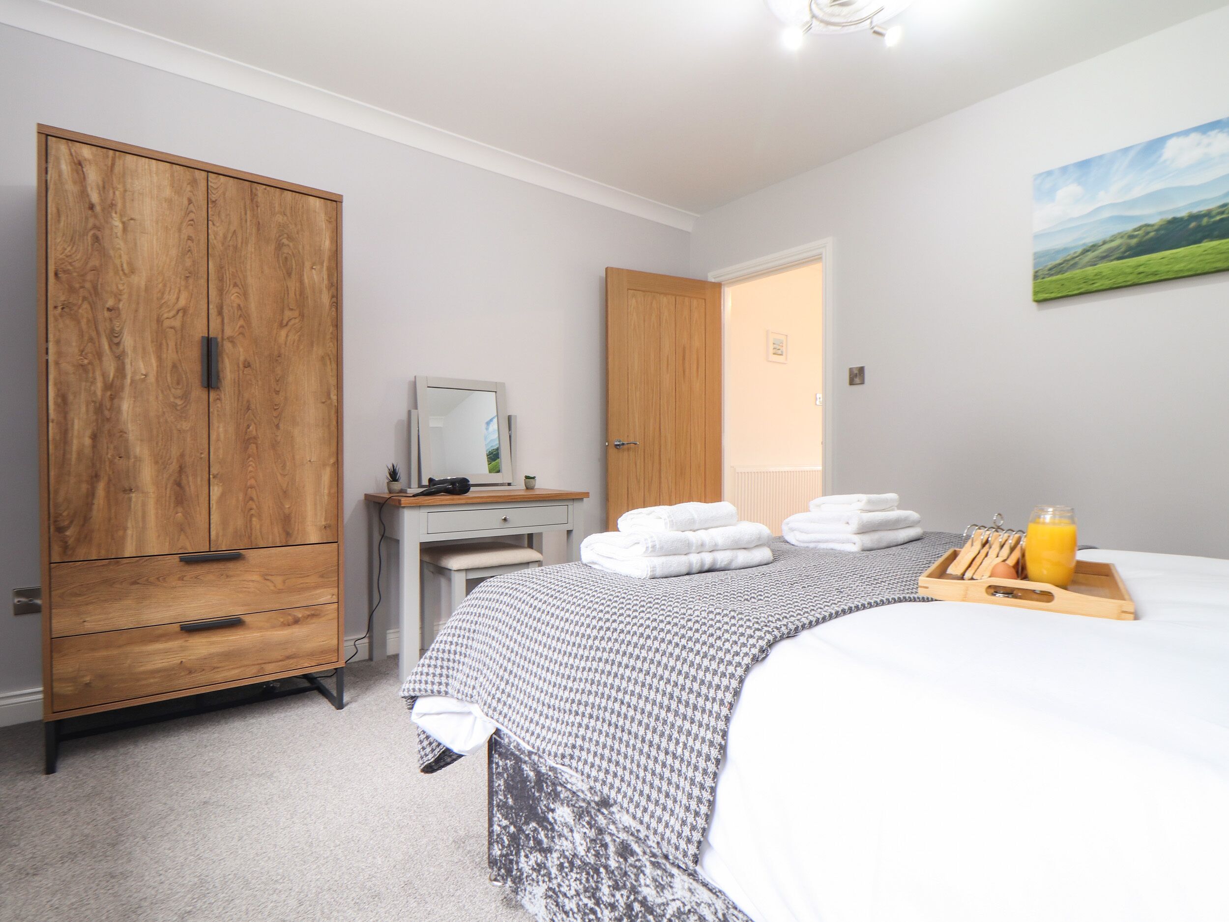 A bedroom with a large wardrobe, a bedside table with a mirror, a bed with a patterned blanket, and a tray with towels and a glass of orange juice.