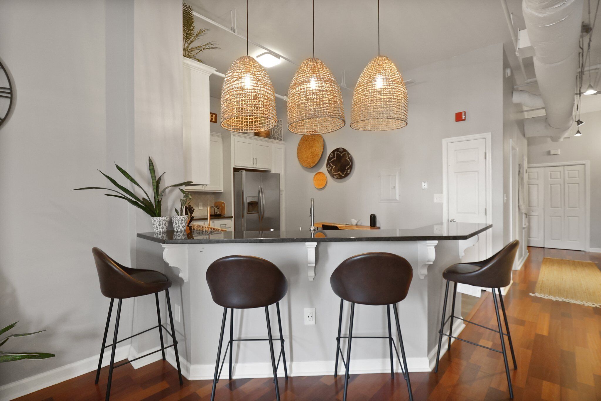 Une cuisine dotée d’un îlot central, de trois tabourets de bar, de luminaires suspendus en osier et d’une plante en pot.