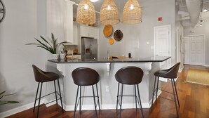 A kitchen with a central island, three bar stools, hanging wicker pendant lights, and a potted plant.
