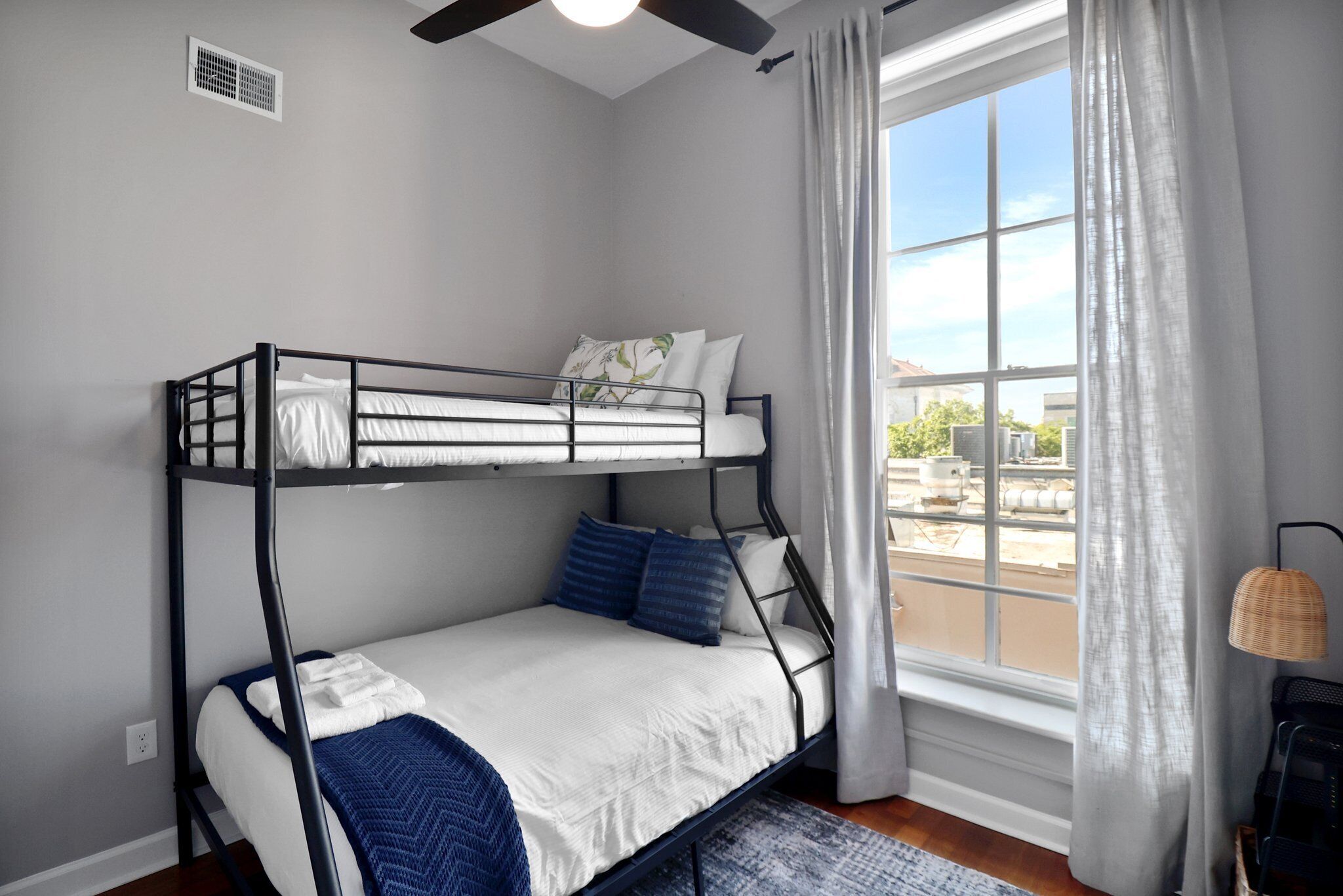 Une chambre avec un lit superposé, une fenêtre, un ventilateur de plafond et une petite table avec une lampe.