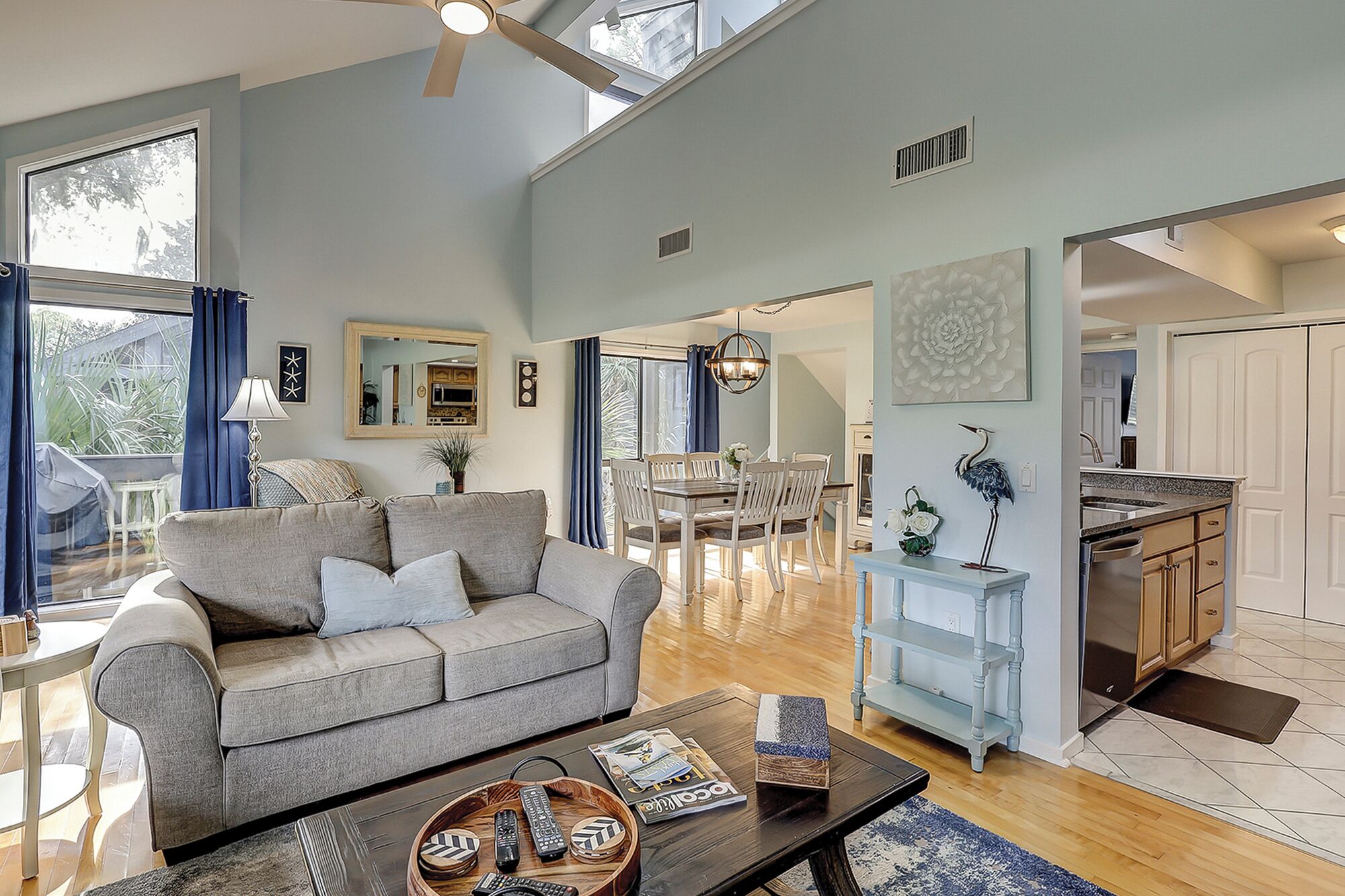 A spacious living room with a grey sofa, a coffee table with magazines, and a dining area with a chandelier.