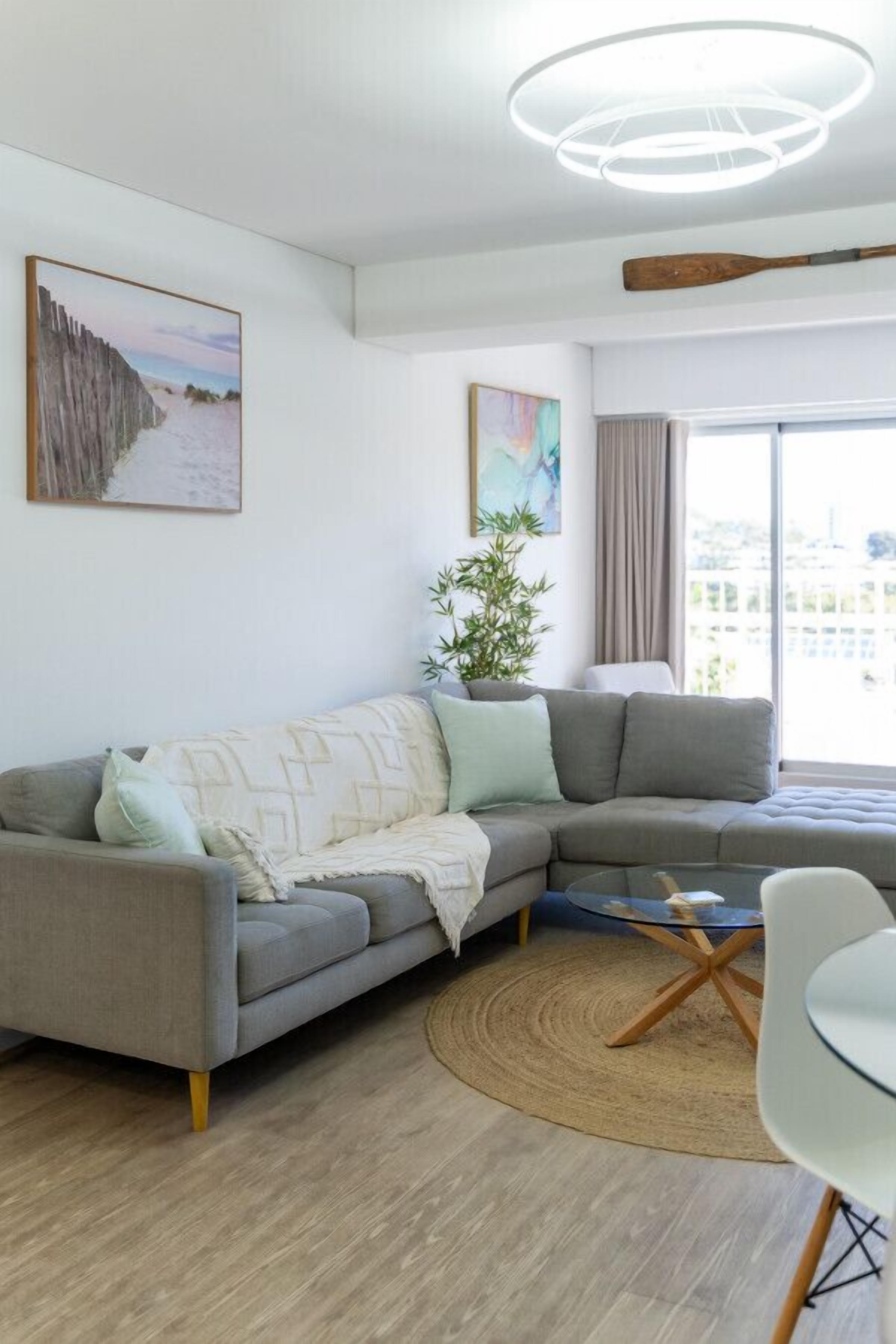 Uma sala de estar moderna com um sofá modular cinza, uma mesa de centro de vidro e um tapete redondo. Há uma janela grande com vista para a cidade e um morcego de madeira na parede.