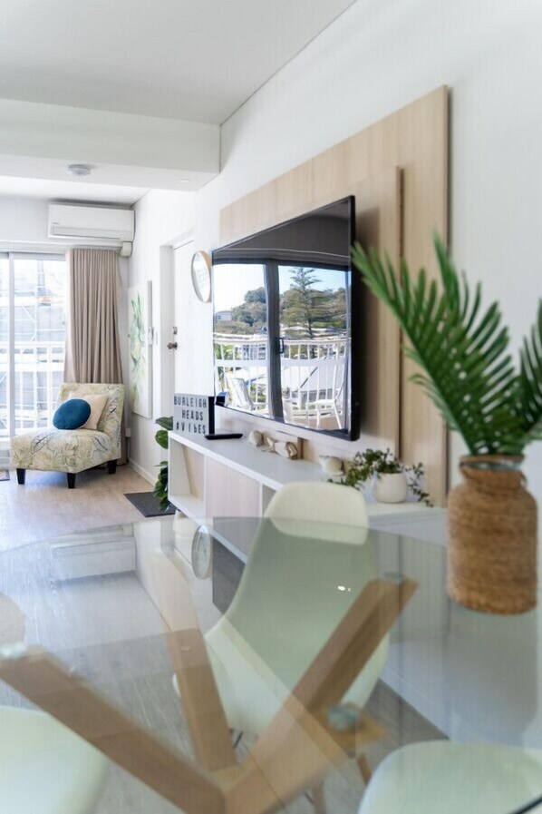 A modern living room with a flat-screen TV mounted on the wall, a comfortable sofa, a glass coffee table, and a view of the outdoors.