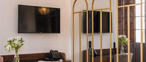 Quarto de hotel moderno com uma grande televisão de tela plana na parede, uma escrivaninha com telefone, uma poltrona confortável e um espelho decorativo com moldura dourada.