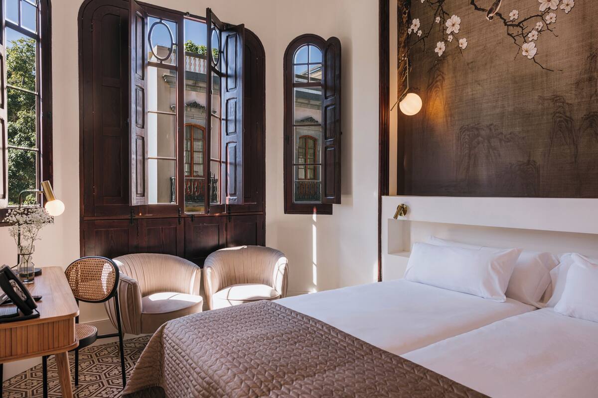 Une chambre d’hôtel avec un grand lit, deux chaises, un bureau avec une lampe et une vue sur un immeuble par la fenêtre.