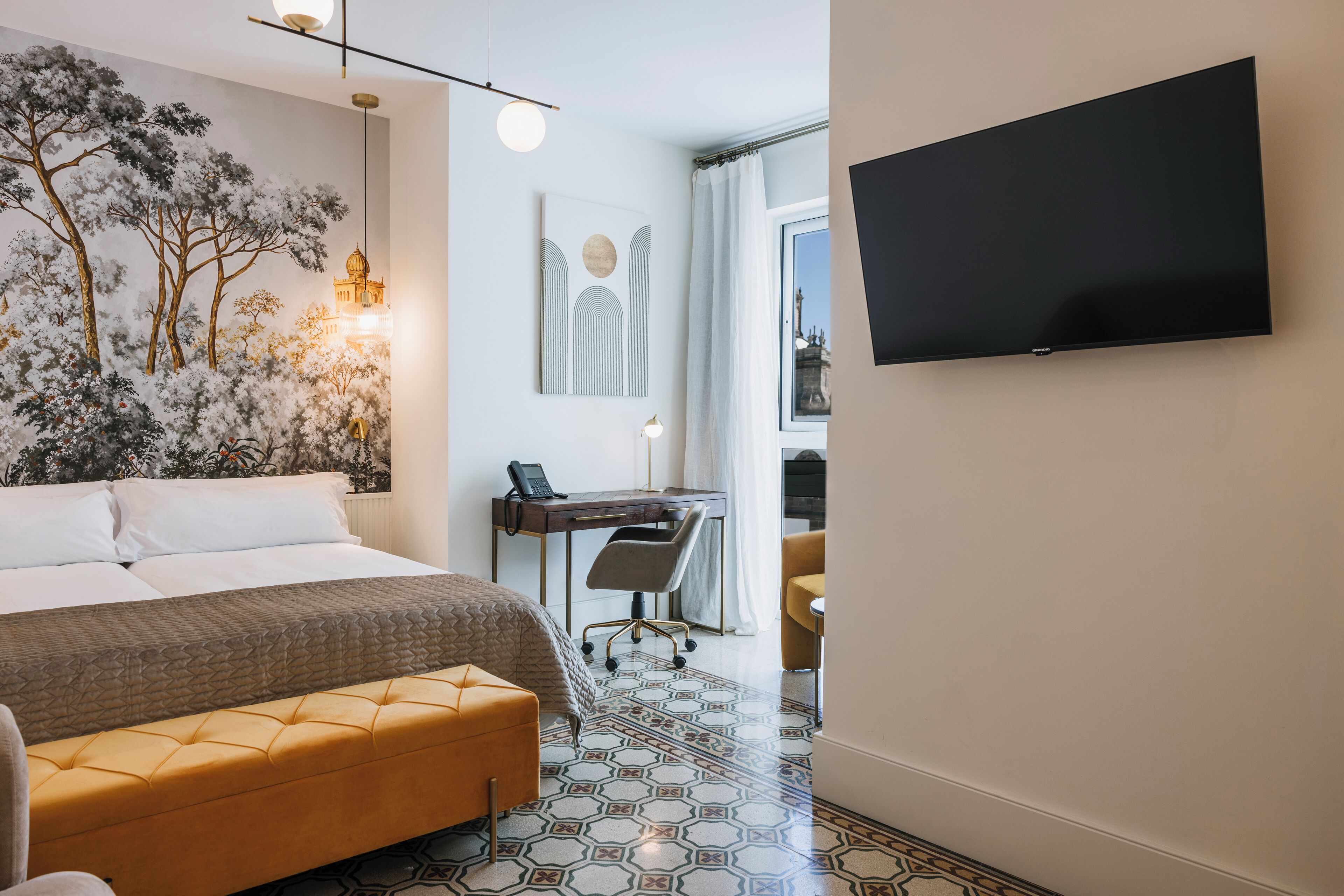 A hotel room with a bed, a desk with a chair, a TV mounted on the wall, and a patterned floor.