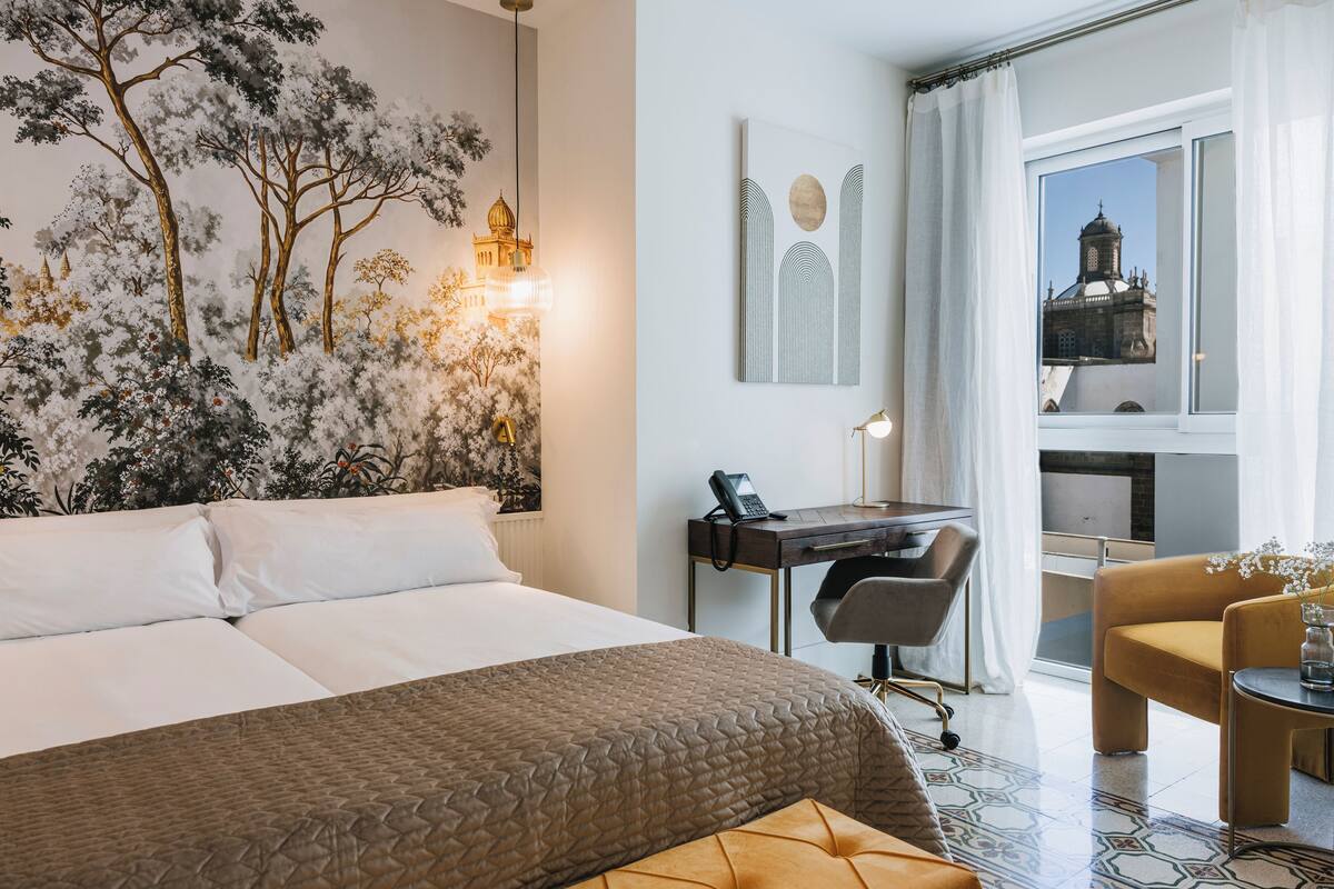 Une chambre d’hôtel avec un grand lit, un bureau avec une chaise, une petite table avec un vase, et une vue sur un immeuble par la fenêtre.