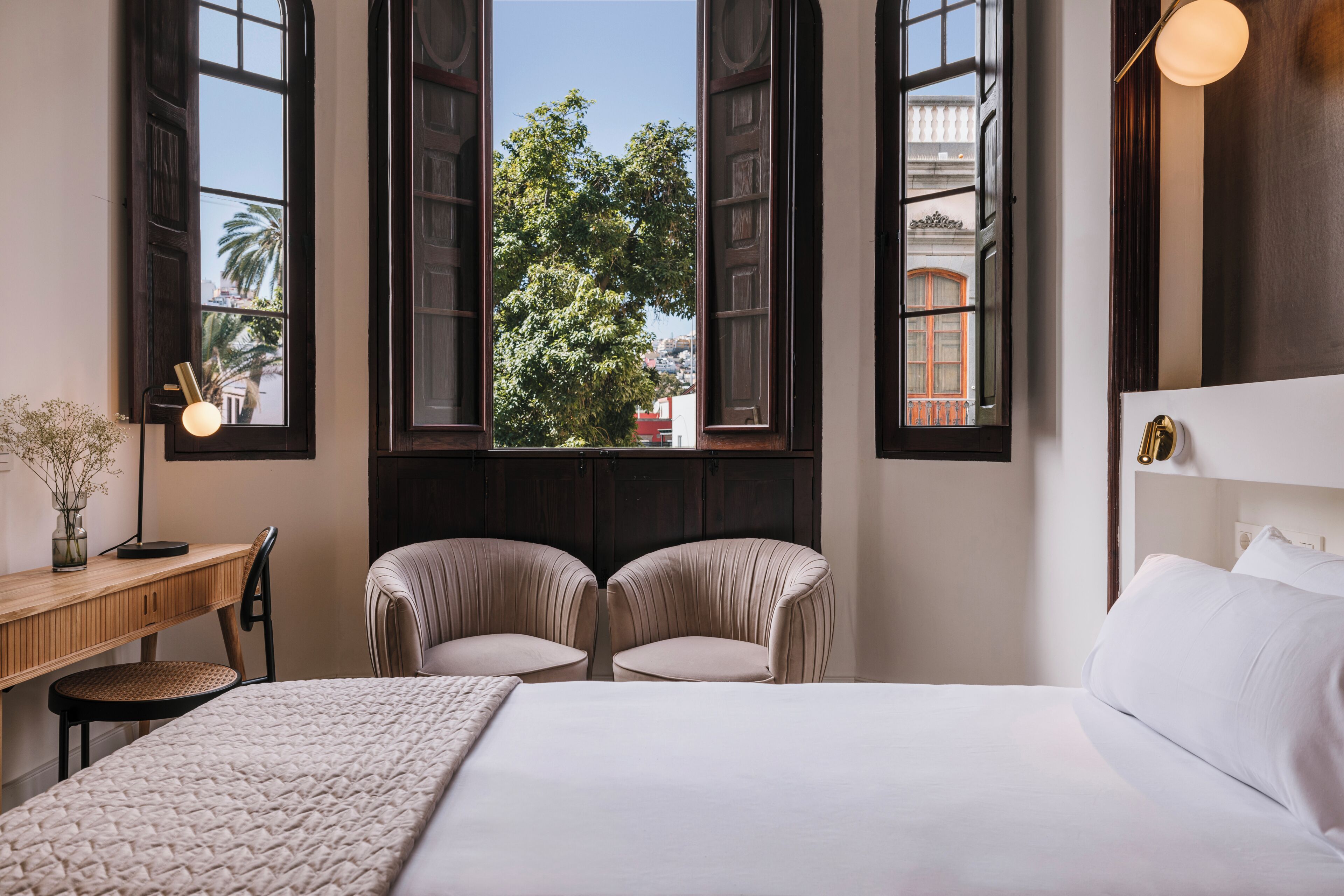 A hotel room with a bed, two chairs, a desk, and large windows with a view of buildings and trees.
