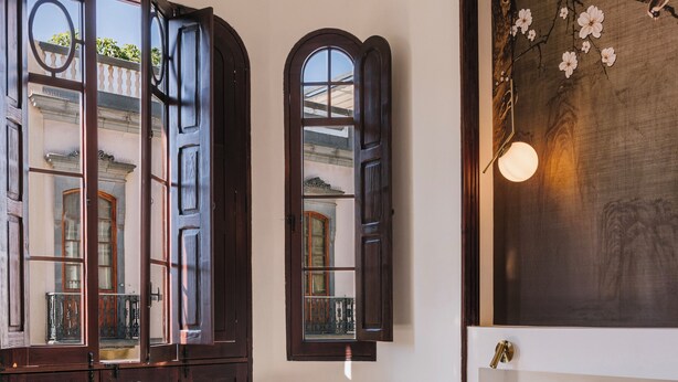 A hotel room with a large bed, two chairs, a wooden floor, and a view of a building through open windows. -  MASANET Emblematic Hotels by Airnest (Las Palmas de Gran Canaria)