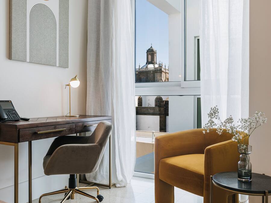 A modern hotel room with a desk, chair, and a view of a historic building through sheer curtains.