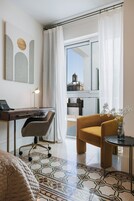 A modern hotel room with a desk, chair, and a view of a historic building through sheer curtains.