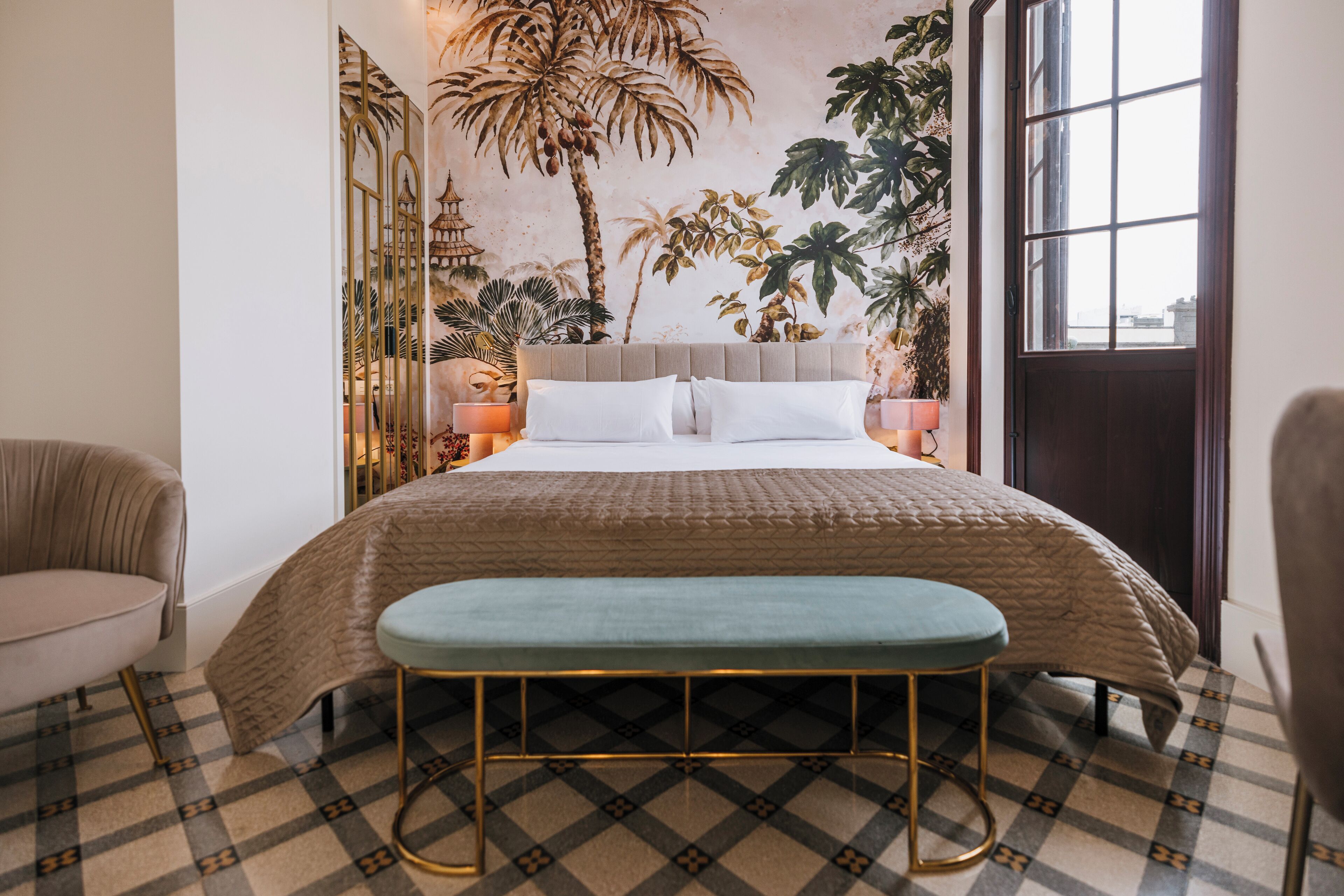 Un dormitorio con una cama grande, un banco, una silla y un mural en la pared con plantas tropicales.