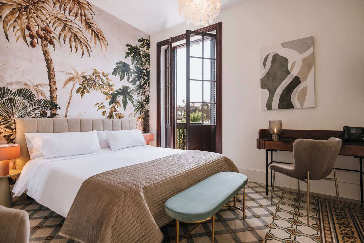 Une chambre d’hôtel avec un lit, un bureau, une chaise, un banc et une grande fenêtre.