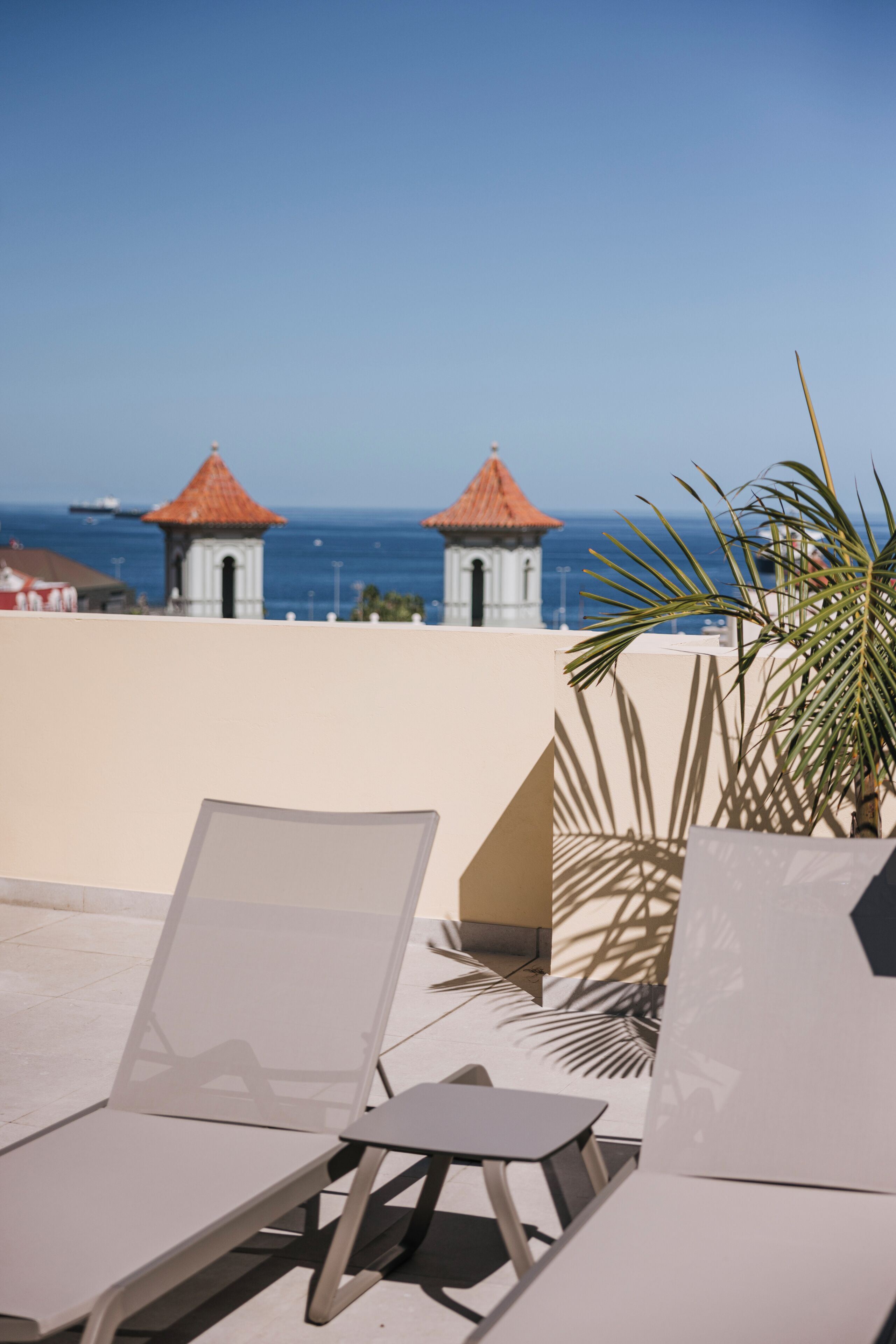 Une terrasse sur le toit, dotée de chaises longues et d’une petite table, offrant une vue sur une zone côtière avec des bâtiments et la mer.
