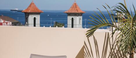 A rooftop terrace with lounge chairs and a small table, overlooking a coastal area with buildings and the sea.