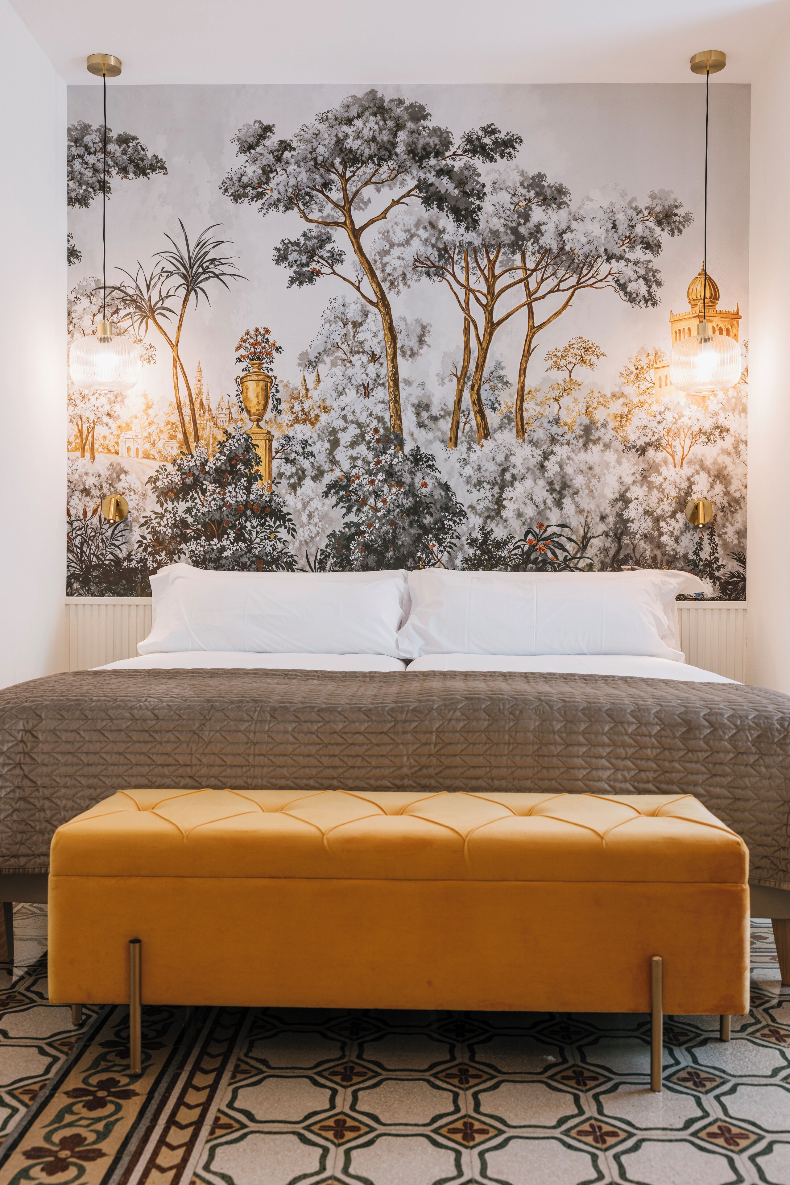 A bedroom with a large bed, a yellow ottoman, and a mural of trees and buildings on the wall.