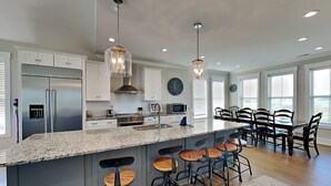 A modern kitchen with a central island, bar stools, and a dining area with a table and chairs. - Family Tides | Spacious Oceanfront Home, Sound Views + Hot Tub! (North Topsail Beach)