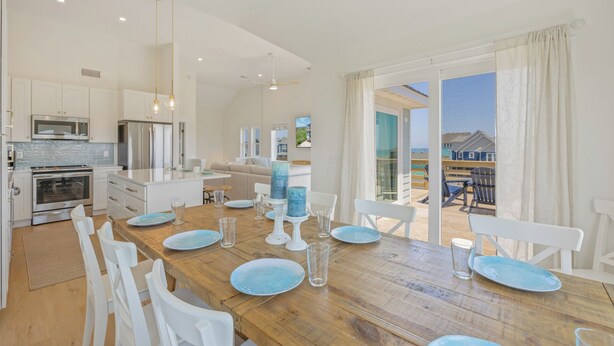 A modern kitchen with stainless steel appliances, a dining area with a wooden table and white chairs, and a view of the beach through large sliding glass doors. - Oceanside Home in Rodanthe with Hot Tub, EV Charging, and Media Rooms (Rodanthe)