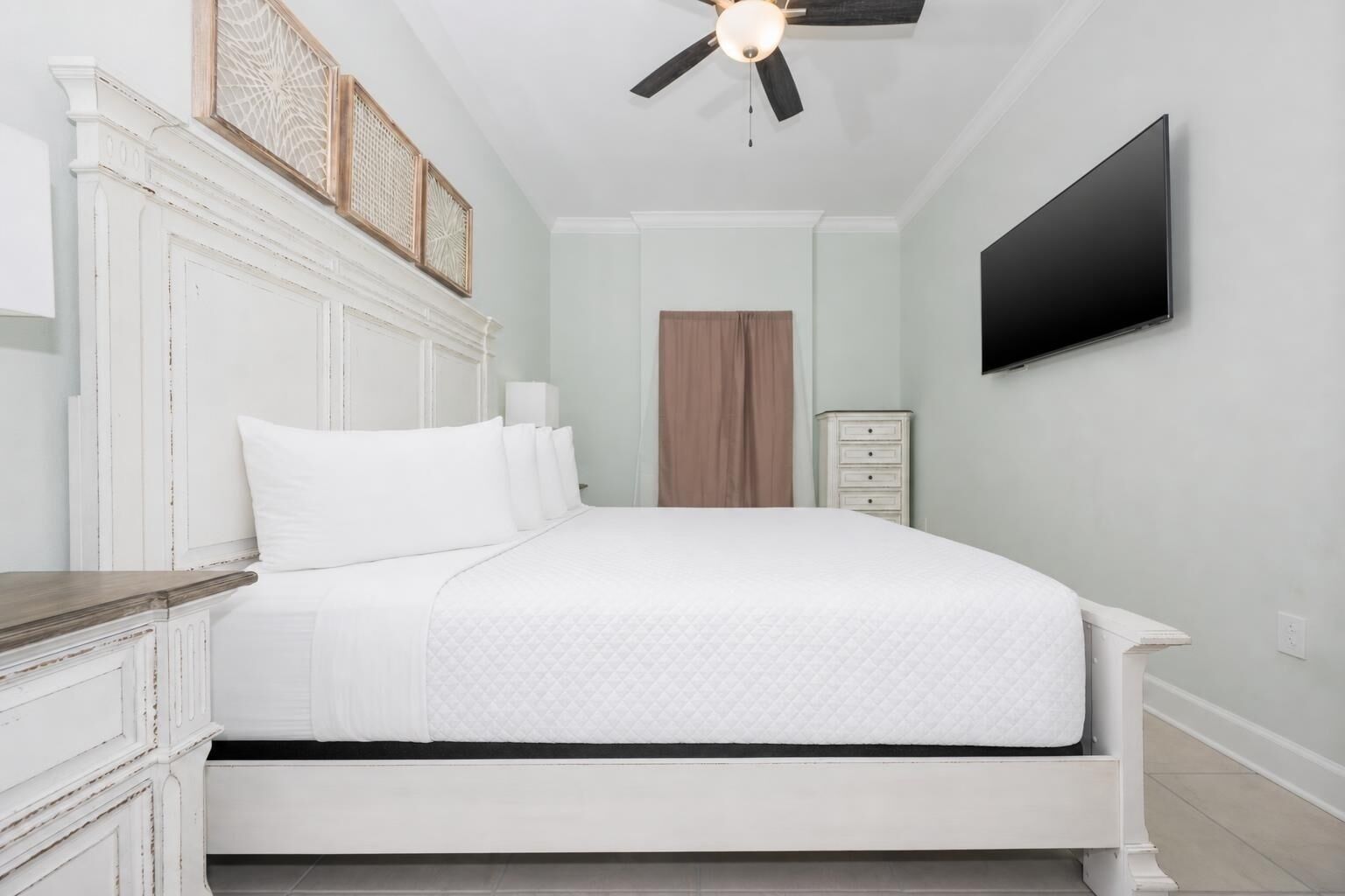 Un dormitorio con una cama, ventilador de techo, televisor montado en la pared y una puerta al fondo.
