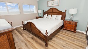 A bedroom with a wooden bed, two bedside tables with lamps, a chair, and a view of the ocean through two windows.