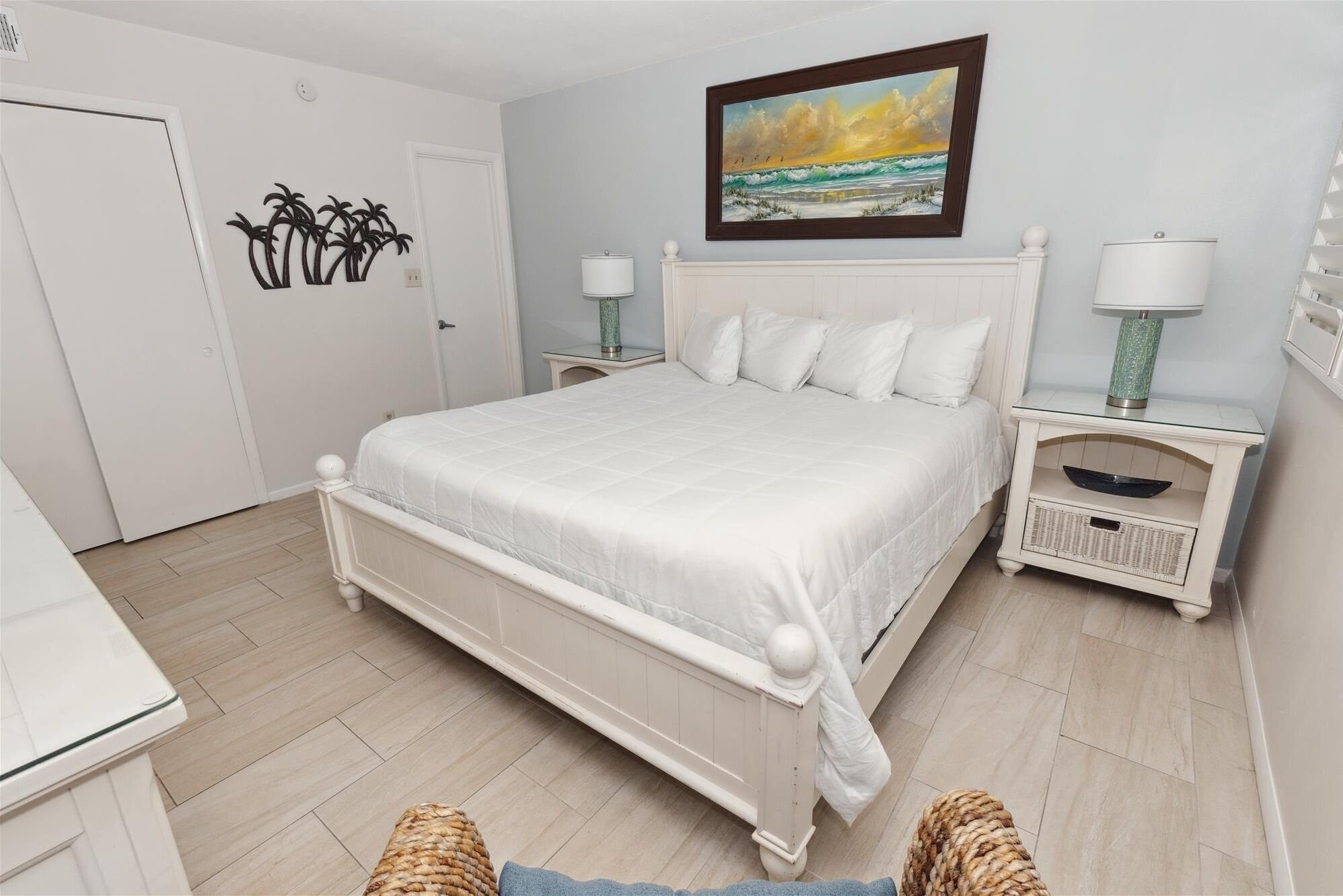 A bedroom with a white bed, two bedside tables with lamps, a painting of a beach scene, and a door with a palm tree decoration.