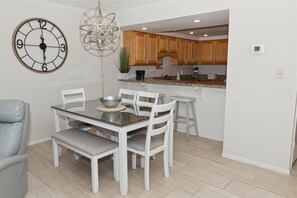 A dining area with a round table, white chairs, and a wall clock. - Beach House 402d 2 Bedroom Condo by RedAwning (Miramar Beach)