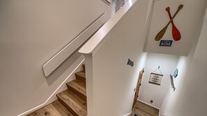 A staircase with wooden steps and white walls, featuring paddleboards hanging on the wall and a sign that says "Y'all come back blinky."
