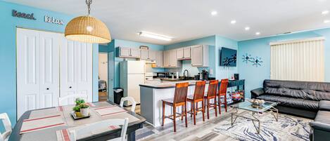 Een moderne woonkamer met een zwarte leren bank, een eethoek met een tafel en stoelen, en een keuken met witte kasten en een centraal keukenblok.