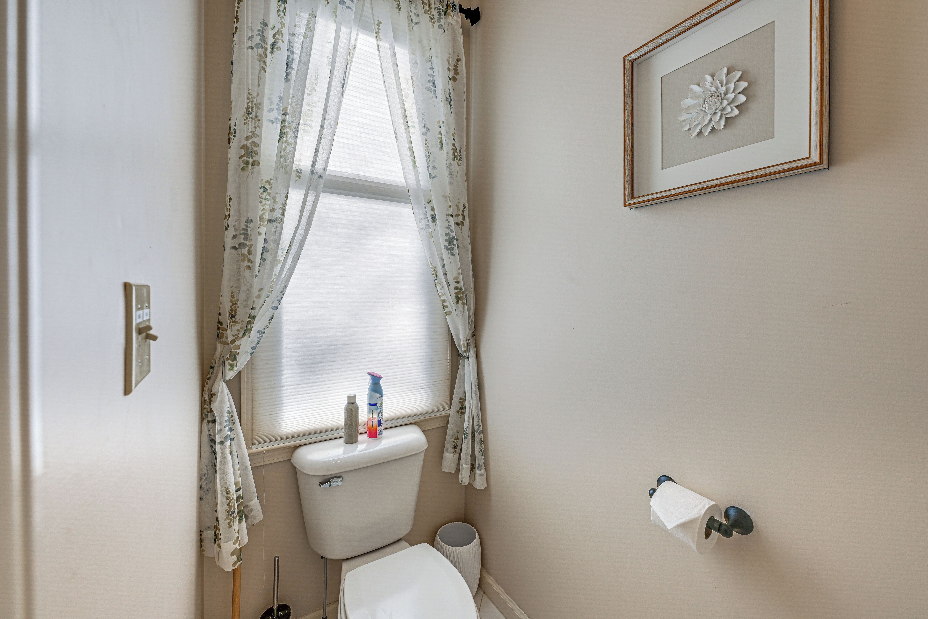 Ein Badezimmer mit Toilette, Waschbecken, einem gerahmten Blumenbild an der Wand und einem Fenster mit floralem Vorhang.