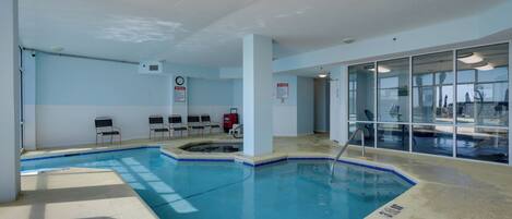 An indoor swimming pool with clear water, surrounded by a tiled floor and walls. There are lounge chairs and a fire extinguisher visible.
