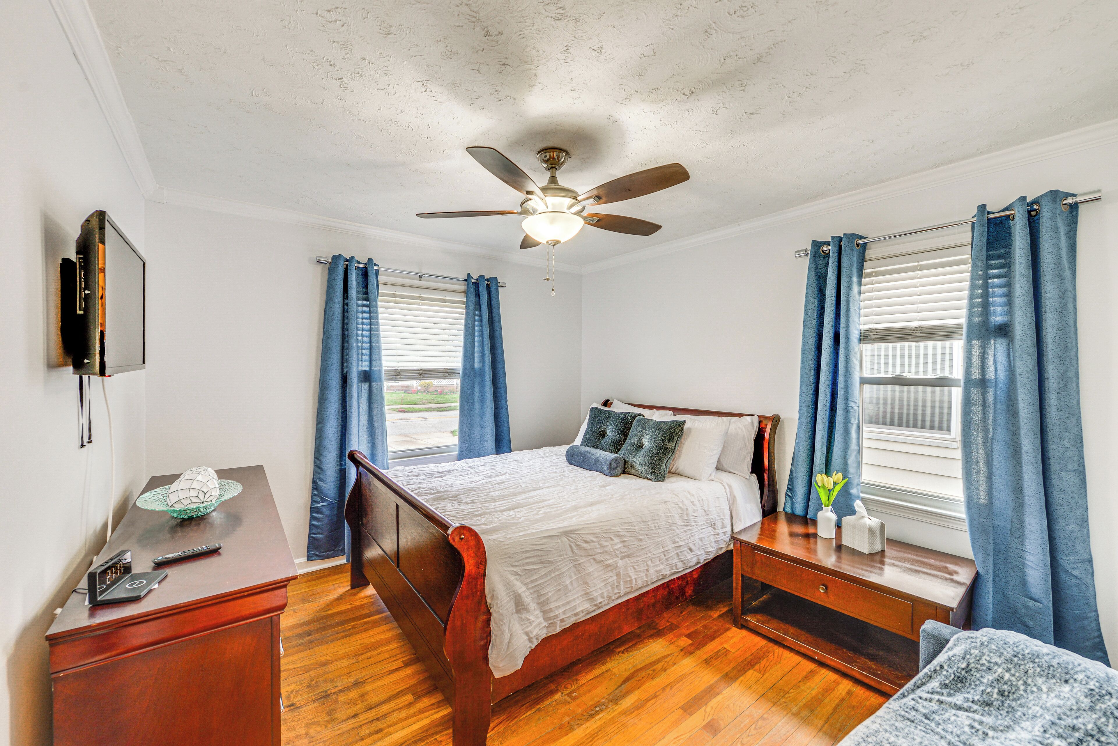 A bedroom with a wooden bed, a ceiling fan, a bedside table, and a TV mounted on the wall.