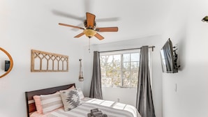 Ein Schlafzimmer mit einem Bett, einem Deckenventilator, einem Fenster mit Vorhängen, einem an der Wand montierten Fernseher und einer Wand-Gitarre.