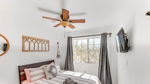 A bedroom with a bed, a ceiling fan, a window with curtains, a mounted TV, and a wall-mounted guitar. - Quiet Oasis w/ Hot Tub: 21 Mi to Joshua Tree! (Landers)