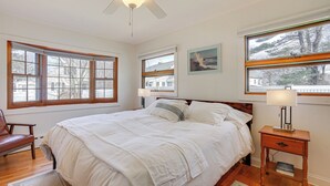 A bedroom with a large bed, wooden headboard, two bedside tables with lamps, a ceiling fan, and large windows with curtains.