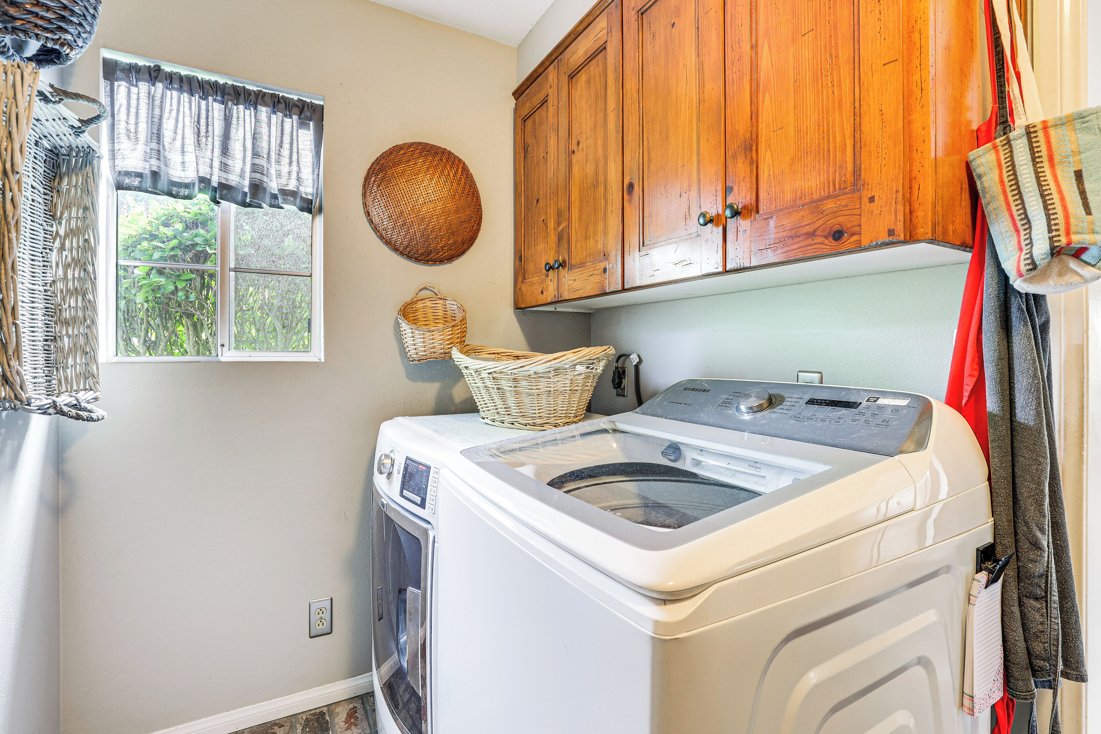 A laundry room with a front-loading washing machine, wooden cabinets, a window with a view of greenery, and a basket on the machine.