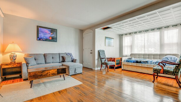 Ein geräumiges Wohnzimmer mit Holzdielenboden, einer Couch, einem Sessel und einem an der Wand montierten Fernseher.