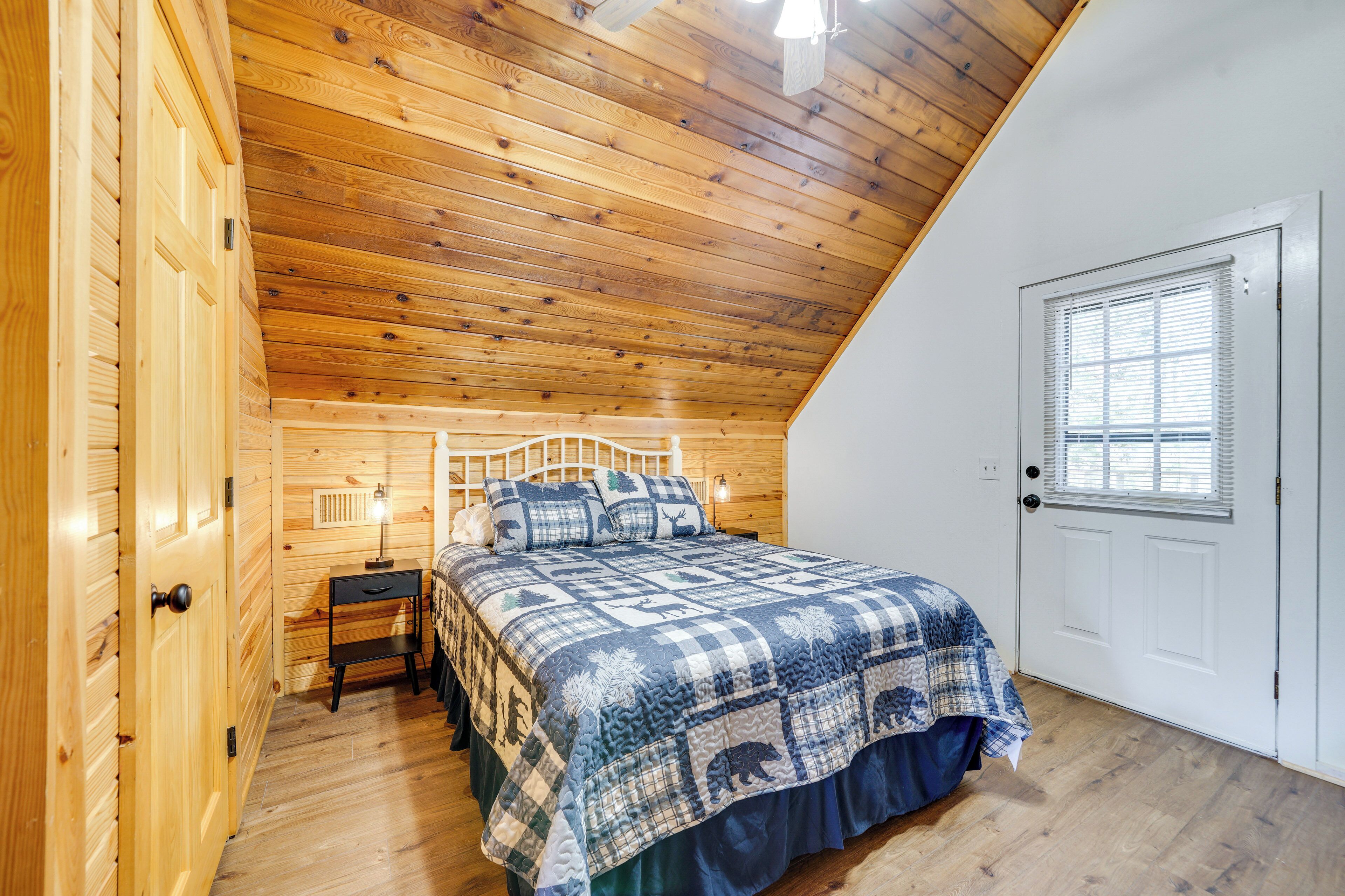 A bedroom with a wooden ceiling, a bed with a blue and white checkered bedspread, a nightstand, a lamp, and a door with a window.