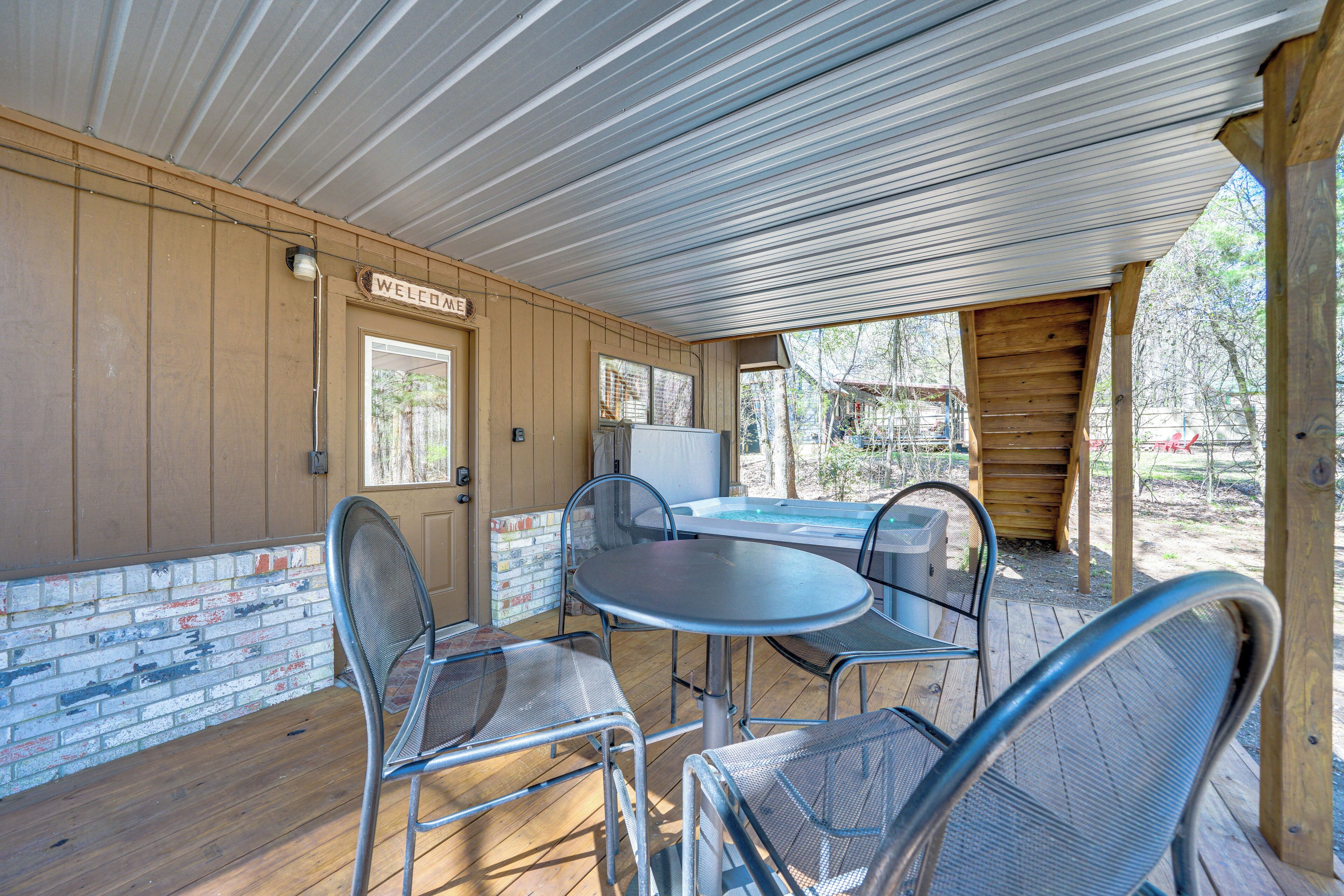 A covered patio with metal chairs and a table, a wooden door with a 'Welcome' sign, and a view of a hot tub and trees.