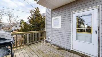 A wooden deck with a grill, a white door with a glass panel, and a gray house with a white trim.