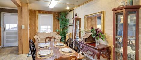A dining area with a wooden table set for four, surrounded by chairs. There is a ceiling fan, a potted plant, and a cabinet with glass doors displaying items.