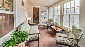 A sunlit room with a wooden floor, white walls, and large windows. There are two metal-framed chairs, a sofa, and a small table with a plant. - Historical 1835 Home in Grand Coteau! (Sunset)