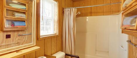 A wooden bathroom with a toilet, sink, and shower. There are shelves with towels and a window with blinds.