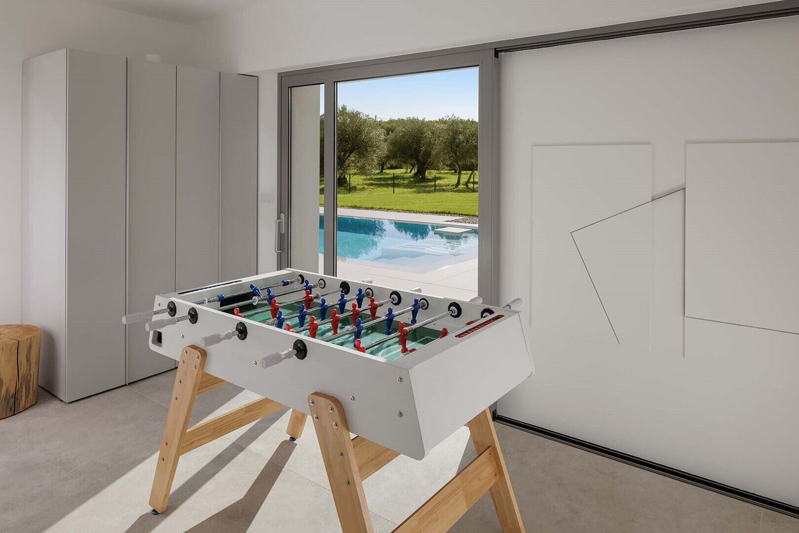 A modern room with a foosball table, a large window with a view of a swimming pool and greenery, and a minimalist design.