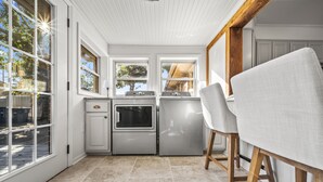 A kitchen with white cabinetry, a built-in oven, and a dishwasher. There are two windows providing natural light, and a wooden chair is visible. - 3BR/3BA 1940s Bungalow Near Expo Square. Pet friendly! (Tulsa)